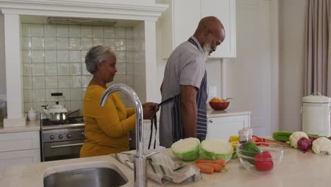 African-american-senior-woman-tying-apron-from-back-to-her-husband-in-the-kitchen-at-home
