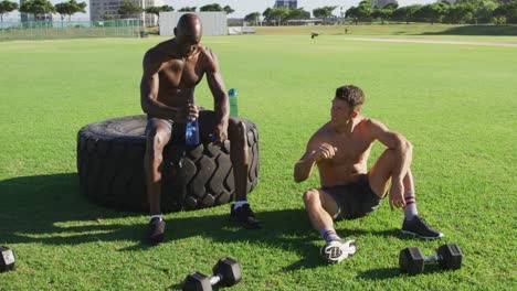 Two-fit,-shirtless-diverse-men-resting,-drinking-water-and-bumping-fists-after-exercising-outdoors