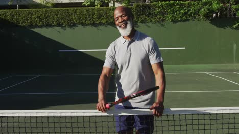 Porträt-Eines-Afroamerikanischen-älteren-Mannes-Mit-Lächelndem-Schläger,-Während-Er-Auf-Dem-Tennisplatz-Steht