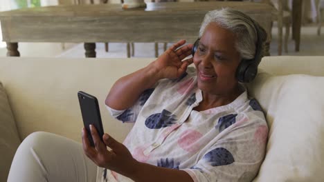 Lächelnde-Afroamerikanische-Seniorin-Mit-Kopfhörern,-Die-Zu-Hause-Einen-Videoanruf-Auf-Dem-Smartphone-Führt