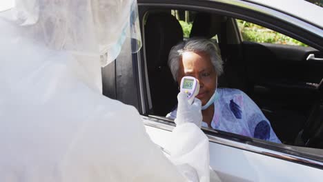 Health-worker-in-protective-clothes-measuring-temperature-of-african-american-senior-woman-in-car