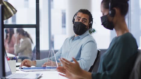 Diverse-male-and-female-colleague-in-face-masks-and-headsets-talking-at-desk-with-sneeze-shield
