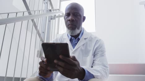 Afroamerikanischer-Männlicher-Arzt,-Der-Im-Krankenhaus-Auf-Der-Treppe-Sitzt-Und-Ein-Tablet-Benutzt