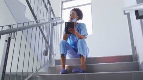 Mixed-race-female-doctor-wearing-face-mask-sitting-on-stairs-in-hospital-using-tablet