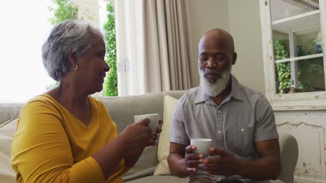 Afroamerikanisches-Seniorenpaar-Lächelt,-Während-Es-Zu-Hause-Auf-Der-Couch-Kaffee-Trinkt