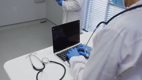 Midsection-of-doctor-wearing-face-mask-and-surgical-gloves-using-laptop,-copy-space-on-screen