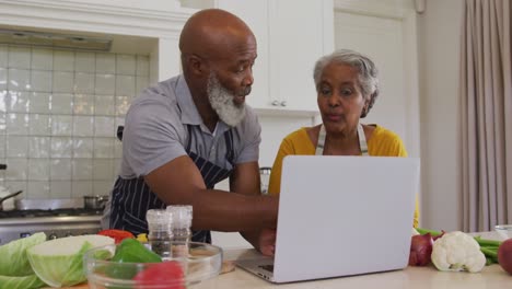 Afroamerikanisches-älteres-Paar-Mit-Schürzen,-Das-Zu-Hause-In-Der-Küche-Einen-Videoanruf-Auf-Dem-Laptop-Führt