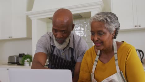 Afroamerikanisches-älteres-Paar-Mit-Schürzen,-Das-Zu-Hause-In-Der-Küche-Einen-Videoanruf-Auf-Dem-Laptop-Führt