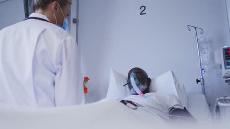 Verschiedener-Arzt-Mit-Gesichtsmaske-Sitzt-Auf-Dem-Krankenhausbett,-Während-Der-Patient-Eine-Sauerstoffmaske-Trägt