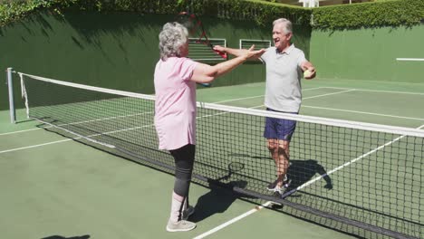 Glückliches-Kaukasisches-älteres-Paar,-Das-Sich-Nach-Einem-Spiel-Auf-Dem-Tennisplatz-Im-Freien-über-Das-Netz-Umarmt