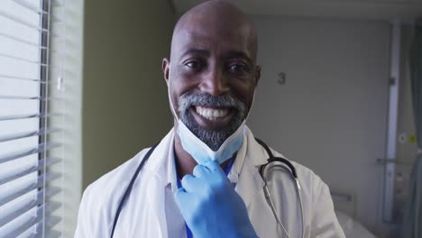 Portrait-of-smiling-african-american-male-doctor-wearing-face-mask-standing-in-hospital-room