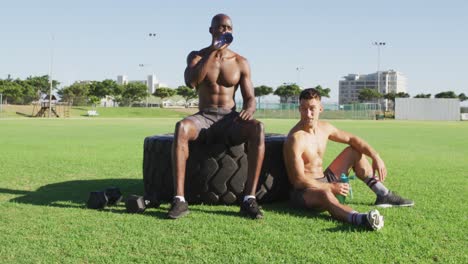 Two-fit,-shirtless-diverse-men-cross-resting,-drinking-water-after-exercising-outdoors