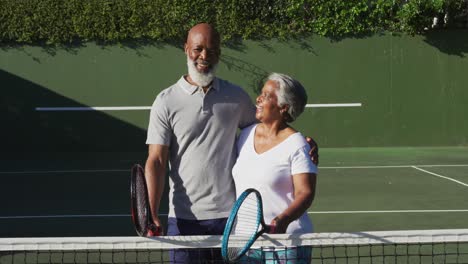 Porträt-Eines-Afroamerikanischen-Seniorenpaares-Mit-Schlägern,-Das-Auf-Dem-Tennisplatz-Steht