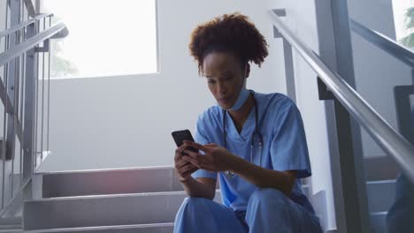 Mixed-race-female-doctor-wearing-face-mask-sitting-on-stairs-in-hospital-using-smartphone