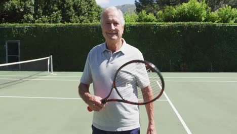 Porträt-Eines-Lächelnden-älteren-Kaukasischen-Mannes,-Der-An-Einem-Sonnigen-Tag-Einen-Tennisschläger-Auf-Dem-Tennisplatz-Hält