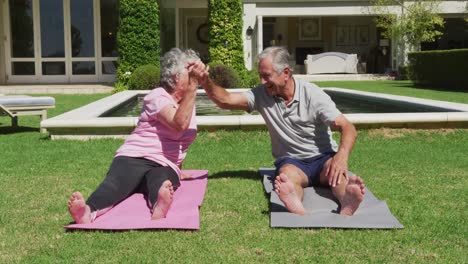 Glückliches-Kaukasisches-älteres-Paar,-Das-Im-Garten-In-Der-Sonne-Yoga-Praktiziert-Und-High-Fiving-Macht
