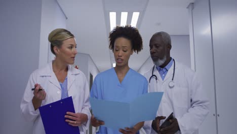 Diverse-male-and-female-doctors-and-medical-staff-walking-hospital-corridor-looking-at-documents