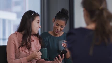 Diverse-group-of-three-happy-businesswomen-drinking-coffee,looking-at-smartphone-and-talking-at-work