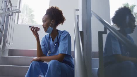 Mixed-race-female-doctor-wearing-face-mask-sitting-on-stairs-in-hospital-talking-on-smartphone