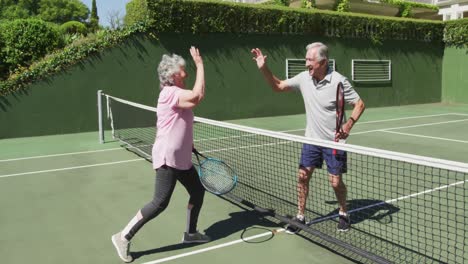 Glückliches-Kaukasisches-Seniorenpaar,-Das-Nach-Einem-Spiel-Auf-Dem-Tennisplatz-Im-Freien-High-Five-über-Das-Netz-Macht