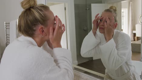 Caucasian-woman-in-bathrobe-touching-her-face-while-looking-in-the-mirror-in-the-bathroom