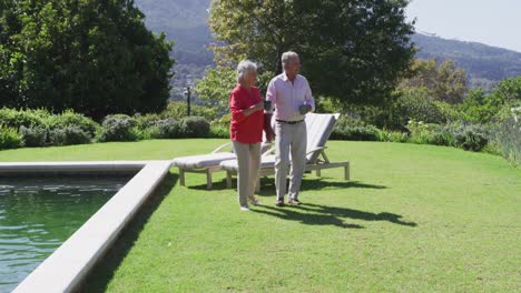 Happy-caucasian-senior-couple-walking-in-sunny-garden-holding-hands-carrying-cups-and-talking
