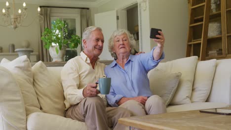 Lächelndes-Kaukasisches-älteres-Paar,-Das-Zu-Hause-Auf-Der-Couch-Sitzt-Und-Ein-Selfie-Vom-Smartphone-Macht