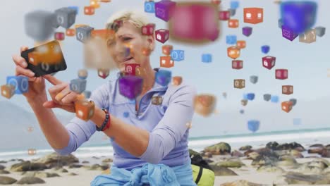 Animation-of-digital-online-icons-over-senior-woman-by-seaside-taking-selfie-with-smartphone