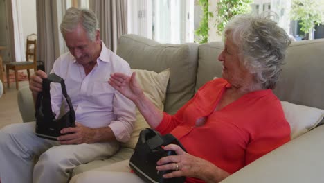 Glückliches-Kaukasisches-Seniorenpaar-Sitzt-Auf-Der-Couch-Im-Wohnzimmer,-Hält-VR-Headsets-In-Der-Hand-Und-Redet