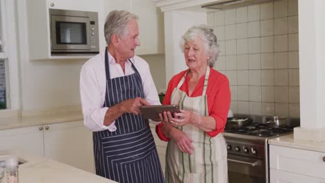 Glückliches-Kaukasisches-älteres-Paar-In-Der-Küche,-Das-Schürzen-Trägt-Und-Vor-Der-Zubereitung-Einer-Mahlzeit-Ein-Tablet-Benutzt