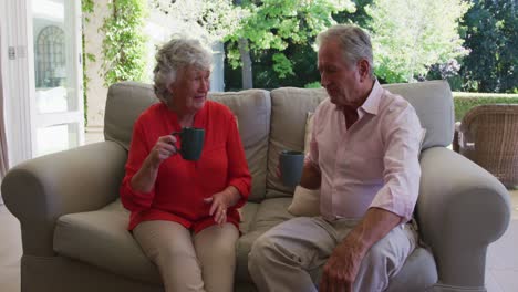 Glückliches-Kaukasisches-Seniorenpaar-Auf-Der-Couch-Im-Sonnigen-Wohnzimmer,-Redet-Und-Trinkt-Eine-Tasse-Kaffee