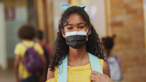 Portrait-of-mixed-race-schoolgirl-wearing-face-mask,-standing-in-corridor-looking-at-camera