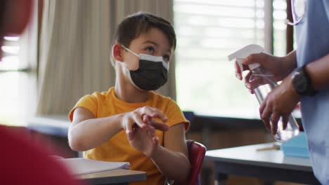 Portrait-of-mixed-race-schoolboy-wearing-face-masks,-disinfecting-hands-in-classroom