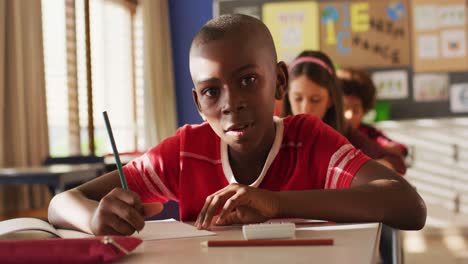 Portrait-of-african-american-schoolboy-sitting-in-class,-making-notes,-looking-at-camera