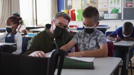 Diverse-male-teacher-helping-schoolboy-sitting-in-classroom,-all-wearing-face-masks