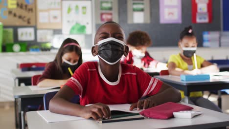Afroamerikanischer-Junge-Trägt-Gesichtsmaske,-Während-Er-In-Der-Klasse-Auf-Seinem-Schreibtisch-Sitzt