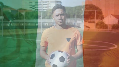 Irish-flag-waving-against-portrait-of-male-soccer-player-holding-soccer-ball-on-grass-field