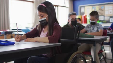 Portrait-of-mixed-race-schoolgirl-sitting-in-wheelchair-in-classroom,-wearing-face-mask
