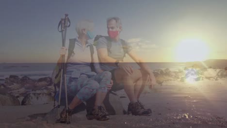 Caucasian-senior-couple-in-face-masks-hiking-on-the-coast-taking-a-rest,-over-moving-sea-at-sunset