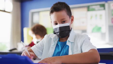Portrait-of-mixed-race-schoolboy-wearing-face-mask-in-classroom-looking-at-camera
