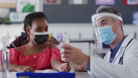Diverse-male-teacher-showing-schoolgirl-how-to-measure-temperature,-all-wearing-face-masks