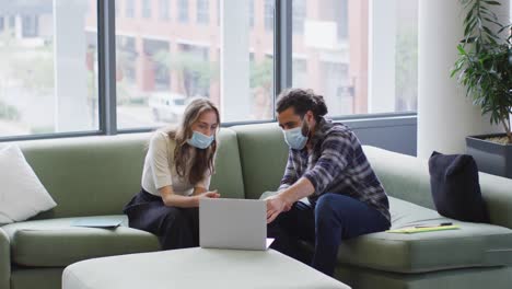 Diverse-male-and-female-business-colleagues-wearing-face-masks-sitting-on-sofa-looking-at-laptop