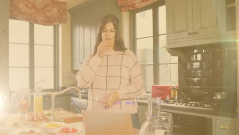 Animation-of-flickering-lights-over-woman-eating-in-kitchen-using-laptop-in-kitchen-at-home