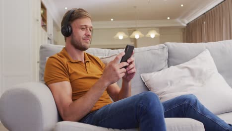 Portrait-of-happy-caucasian-man-sitting-on-sofa-wearing-headphones-and-using-smartphone