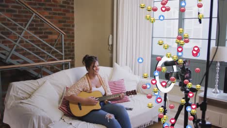Animation-of-emoji,-love-and-like-icons-over-female-vlogger-with-guitar-recording-vlog-at-home