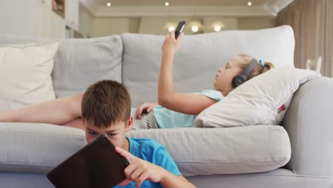 Portrait-of-caucasian-brother-and-sister-lying-on-sofa-using-tablet-and-smartphone