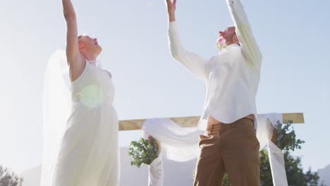 Happy-caucasian-newly-wed-couple,-jumping-throwing-petals-in-front-of-altar-outdoors
