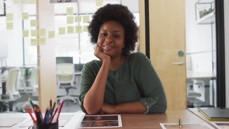 Porträt-Einer-Afroamerikanischen-Geschäftsfrau,-Die-Am-Schreibtisch-Sitzt,-Sich-Auf-Die-Hand-Stützt-Und-Im-Büro-Lächelt