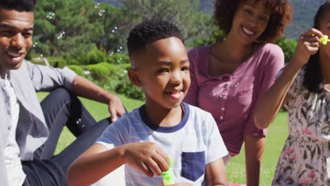Glückliche-Afroamerikanische-Eltern,-Tochter-Und-Sohn-Sitzen-Draußen-Auf-Einer-Decke-Und-Blasen-Seifenblasen