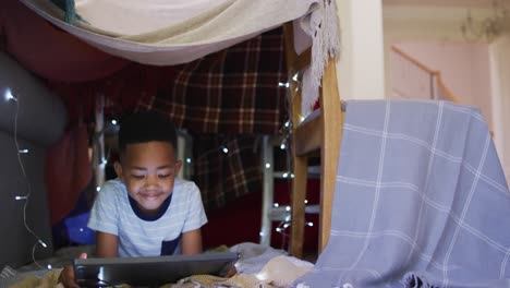 Happy-african-american-boy-lying-in-house-made-of-blankets,-using-tablet